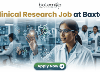 Latest Clinical Research Job for Life Science & Biomedical Graduates at Baxter | Apply Now Latest Clinical Research Job at Baxter | Apply Now. A featured job advertisement banner for biotecnika, titled "Clinical Research Job at Baxter," set in a modern, futuristic laboratory. The visual focuses on a diverse female scientist, of South Asian or Indian descent, with safety glasses and a lab coat, who is interacting with and gesturing towards a floating, transparent holographic screen. The screen displays complex 3D molecular models of a protein and a drug, along with labels like 'Protein Drug Docking' and 'Data Docking'. In the blurred background, other researchers work with equipment and computer monitors in a well-equipped facility. At the top center is the "biotecnika Your Bio Resource" logo. At the bottom center, a prominent green button contains the text 'Apply Now' with a white arrow icon.