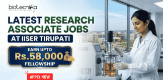 Latest Research Associate Jobs at IISER Tirupati | Earn Upto Rs.58,000 Fellowship Latest Research Associate Jobs at IISER Tirupati | Earn Upto Rs.58,000 Fellowship. A professional recruitment banner for "Latest Research Associate Jobs at IISER Tirupati," featuring the Biotecnika logo at the top left. On the right, a female scientist in a white lab coat and gloves is shown working with a laboratory bioreactor in a bright, modern lab setting. The background through a large window offers a realistic view of the IISER Tirupati campus. The left side of the image features the bold headline "LATEST RESEARCH ASSOCIATE JOBS AT IISER TIRUPATI." Below this, a prominent gold-framed blue box highlights the fellowship: "EARN UPTO Rs. 58,000 FELLOWSHIP" accompanied by an icon of a money bag. Three circular icons highlight key benefits: Conduct Advanced Bio-Research, Collaboration & Mentorship, and Career Growth Path. A large green "Apply Now" button with a white arrow and the website "biotecnika.org" are positioned at the bottom.