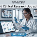 Hybrid Clinical Research Job at IQVIA Opening for Life Science Graduates! A promotional graphic titled "Hybrid Clinical Research Job at IQVIA" at the top center, just below the biotecnika logo. The central image is a female researcher in a lab coat, nitrile gloves, and safety glasses, focused on carefully using a pipette on a multi-well plate. To her left is a large tablet displaying various clinical research charts and data plots. To her right is a paper titled "Remote Connection" and a coffee cup with a sleeve that reads "flex work". Large screens in the background show other scientists collaborating and a multi-person video conference call with a diverse team under the title "Global Team Collaboration". A centrifuge and other lab equipment are visible in the background. A prominent blue button at the bottom center reads "APPLY NOW".