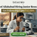 Junior Research Fellow Job at University of Allahabad | Get Fellowship Upto ₹42,000 Junior Research Fellow Job at University of Allahabad Get Fellowship Upto ₹42,000. A job advertisement graphic for University of Allahabad. The top has text: 'University of Allahabad Hiring Junior Research Fellow' and 'Get Fellowship Upto ₹42,000.' A central photo shows a woman in a lab coat using a microscope in a laboratory, with an old Indian university building visible through a window. A desk plaque reads 'NAM. SURIANDAH (GENERIC JRF)' and other lab equipment is present. A prominent green 'APPLY NOW' button is at the bottom center. The biotecnika logo is at the top center.