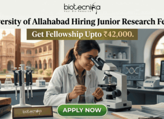Junior Research Fellow Job at University of Allahabad Get Fellowship Upto ₹42,000. A job advertisement graphic for University of Allahabad. The top has text: 'University of Allahabad Hiring Junior Research Fellow' and 'Get Fellowship Upto ₹42,000.' A central photo shows a woman in a lab coat using a microscope in a laboratory, with an old Indian university building visible through a window. A desk plaque reads 'NAM. SURIANDAH (GENERIC JRF)' and other lab equipment is present. A prominent green 'APPLY NOW' button is at the bottom center. The biotecnika logo is at the top center.