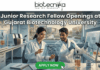 Junior Research Fellow at Gujarat Biotechnology University | 36K/ Month Fellowship Junior Research Fellow Openings at Gujarat Biotechnology University 36,000 Month Fellowship. A promotional banner for Junior Research Fellow openings at Gujarat Biotechnology University, presented by Biotecnika. Two smiling scientists in white lab coats are shown in a futuristic laboratory. The female scientist points to a holographic display featuring a DNA helix and specific details about DNA fingerprinting, genome editing success, DNA sequencing, research areas, and eligibility (M.Sc/M.Tech). Labeling in the background points to "Genome Engineering" and "Bioprocess Lab," and a coffee mug with "Caffeine & CRISPR" is visible. A prominent green "APPLY NOW" button is at the bottom center with a mouse pointer icon.