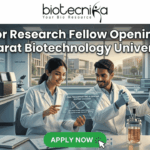 Junior Research Fellow at Gujarat Biotechnology University | 36K/ Month Fellowship Junior Research Fellow Openings at Gujarat Biotechnology University 36,000 Month Fellowship. A promotional banner for Junior Research Fellow openings at Gujarat Biotechnology University, presented by Biotecnika. Two smiling scientists in white lab coats are shown in a futuristic laboratory. The female scientist points to a holographic display featuring a DNA helix and specific details about DNA fingerprinting, genome editing success, DNA sequencing, research areas, and eligibility (M.Sc/M.Tech). Labeling in the background points to "Genome Engineering" and "Bioprocess Lab," and a coffee mug with "Caffeine & CRISPR" is visible. A prominent green "APPLY NOW" button is at the bottom center with a mouse pointer icon.