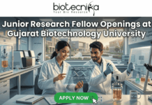 Junior Research Fellow at Gujarat Biotechnology University | 36K/ Month Fellowship Junior Research Fellow Openings at Gujarat Biotechnology University 36,000 Month Fellowship. A promotional banner for Junior Research Fellow openings at Gujarat Biotechnology University, presented by Biotecnika. Two smiling scientists in white lab coats are shown in a futuristic laboratory. The female scientist points to a holographic display featuring a DNA helix and specific details about DNA fingerprinting, genome editing success, DNA sequencing, research areas, and eligibility (M.Sc/M.Tech). Labeling in the background points to "Genome Engineering" and "Bioprocess Lab," and a coffee mug with "Caffeine & CRISPR" is visible. A prominent green "APPLY NOW" button is at the bottom center with a mouse pointer icon.