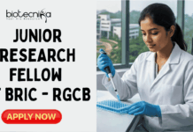 Junior Research Fellow Vacancy 2026 at BRIC – RGCB | Earn ₹37,000 Salary Junior Research Fellow Vacancy 2026 at BRIC – RGCB Earn ₹37,000 Salary. A featured job advertisement image for a Junior Research Fellow position at BRIC-RGCB. At the top center is the biotecnika Your Bio Resource logo. The image features a young woman researcher in a lab coat and gloves, seated at a modern lab bench and carefully pipetting into a microplate. Surrounding her are lab equipment and a monitor showing scientific data. Large glass windows in the background look out onto a modern research campus with greenery. A prominent central text overlay reads, "JUNIOR RESEARCH FELLOW VACANCY at BRIC – RGCB". At the bottom center is a green button that says "APPLY NOW".
