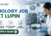 Latest Biology Job At Lupin | BSc Candidates Apply Before It’s Gone! Latest Biology Job At Lupin BSc Candidates Apply Before It's Gone! A detailed, professional recruitment banner for a "BIOLOGY JOB AT LUPIN" by biotecnika Your Bio Resource. The banner features a focused female Indian scientist in a lab coat and safety glasses, using a multichannel pipette to fill a microplate in a modern laboratory. Overlaid on the background, which includes a campus view and scientific monitors with bio-prospecting data and molecular models, are hexagonal panels displaying a DNA helix, a cell structure, and a plant sample. Three key career benefits are listed with icons in green circles: "CUTTING-EDGE DRUG RESEARCH", "DRUG DEVELOPMENT PATHWAYS", and "CAREER ACCELERATION". A prominent green "APPLY NOW" button with a white right arrow is at the bottom center. The company logo is at the top left, and the job title text is prominent on the left.