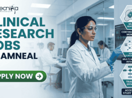 Latest Clinical Research Jobs at Amneal Apply Before It Closes! A professional corporate recruitment banner for "CLINICAL RESEARCH JOBS AT AMNEAL" featuring the Biotecnika logo. The image showcases a focused female scientist of Indian heritage in a white lab coat and safety goggles, interacting with a high-tech digital monitor displaying clinical trial data and ECG waveforms. To the right, hexagonal icons highlight key clinical research themes: "Continuous Patient Monitoring," "Multi-modal Data Integration," and "Accelerated Study Timelines." The background depicts a modern, brightly lit clinical facility with other researchers at work. A prominent green "APPLY NOW" button with a white arrow is positioned at the bottom left.
