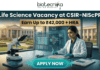 Latest Life Science Vacancy at CSIR-NIScPR | MSc Candidates Earn Upto ₹42,000/- Life Science Vacancy at CSIR-NIScPR Earn Up to ₹42,000 + HRA. A promotional job advertisement banner for a "Life Science Vacancy at CSIR-NIScPR." The central image features a female researcher in a lab coat and gloves at a sophisticated laboratory desk. She is using a micropipette with a futuristic, transparent data display. Foreground objects include a microscope, a multi-well plate, stacks of scientific journals, and a DNA helix model. Behind her are large windows showing a grand, classical-style campus building under a sunny sky, representing the institute. At the top center, the Biotecnika "Your Bio Resource" logo is visible. The text reads: "Life Science Vacancy at CSIR-NIScPR", with a prominent gold banner below highlighting: "Earn Up to ₹42,000 + HRA". A large, pulsing teal button at the bottom center says "APPLY NOW."