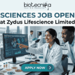 Life Sciences Job Openings at Zydus Lifescience Limited Walk-In Interview 2026. A promotional banner from Biotecnika, titled 'LIFE SCIENCES JOB OPENINGS at Zydus Lifescience Limited'. The image features a photo of three diverse and collaborative researchers in a modern laboratory. One woman in a lab coat is smiling and pointing to a computer monitor displaying 'SUCCESSFUL PHASE III RESULTS' and scientific data visualizations. Her colleagues look on with interest. Laboratory tools like pipettes, petri dishes with samples, vials, and a laptop are visible on the desk. At the bottom center, a prominent blue button reads 'APPLY NOW' with an arrow icon. The overall theme is successful, modern scientific collaboration.