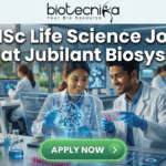 MSc Life Science Job Opening at Jubilant Biosys Apply Online Today! A featured job recruitment banner with the text "MSc Life Science Job at Jubilant Biosys" positioned prominently below the Biotecnika logo. The central image is a photorealistic illustration of a diverse male and female scientist pair in lab coats collaborating in a modern, sunlit laboratory. They are interacting with a futuristic holographic screen that displays a complex 3D molecular structure, accompanied by smaller text details like "Target Protein-Ligand Affinity" and "Lead Compound Screen Results". One scientist holds a microplate with colorful well contents, and the other points at the holographic display. A large, green, prominent "APPLY NOW" button is at the bottom center. The lab background features advanced scientific equipment and a view of a park through a large window.