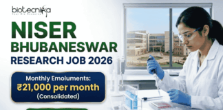 NISER Bhubaneswar Research Job 2026 – Life Sciences Graduates Can Apply NISER Bhubaneswar Research Job 2026 – Life Sciences Graduates Can Apply. A professional recruitment banner for "NISER Bhubaneswar Research Job 2026," featuring the Biotecnika logo at the top left. On the right, a female scientist in a white lab coat and blue gloves is precisely using a pipette on a multi-well plate in a bright, modern laboratory. The left side displays the bold headline "NISER BHUBANESWAR RESEARCH JOB 2026." Below the title, a prominent gold-framed blue box highlights the salary: "Monthly Emoluments: ₹21,000 per month (Consolidated)." The background through the lab window shows a realistic view of the NISER Bhubaneswar campus. A large green "Apply Now" button with a white arrow is positioned at the bottom left.