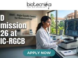 PhD Admission 2026 at BRIC-RGCB Unlock Your Life Science Career Dream! Promotional banner for Biotecnika, "Your Bio Resource," advertising PhD Admission 2026 at BRIC-RGCB (Rajiv Gandhi Centre for Biotechnology). The central image is of a female scientist in safety glasses and a lab coat working with advanced lab equipment and a large computer monitor displaying DNA and protein visualizations. In the background, a sign on a glass partition reads "BRIC - Rajiv Gandhi Centre for Biotechnology," and a modern research campus is visible through windows. At the bottom, a prominent green button with white text says "APPLY NOW."