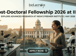 Post-Doctoral Fellowship At IISc Bengaluru Earn ₹2 Lakh Grant. An attractive and realistic featured image for a job page from biotecnika. At the top, the biotecnika logo is centered. Large, bold centered text below reads, "Post-Doctoral Fellowship 2026 at IISc," with a smaller sub-headline that says, "EXPLORE ADVANCED RESEARCH AT INDIA'S PREMIER INSTITUTE | MAY 2026." The image features a female Indian researcher smiling in a modern laboratory, holding documents. Various lab equipment like microscopes and other scientists in the background are visible. Through the large windows, a recognizable, grand academic building and trees are visible, implying the IISc campus under a warm, golden light. At the bottom center, a rounded button reads "APPLY NOW >." The image is relatable, attention-grabbing, and unique.