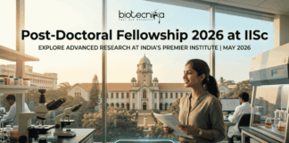Post-Doctoral Fellowship At IISc Bengaluru Earn ₹2 Lakh Grant. An attractive and realistic featured image for a job page from biotecnika. At the top, the biotecnika logo is centered. Large, bold centered text below reads, "Post-Doctoral Fellowship 2026 at IISc," with a smaller sub-headline that says, "EXPLORE ADVANCED RESEARCH AT INDIA'S PREMIER INSTITUTE | MAY 2026." The image features a female Indian researcher smiling in a modern laboratory, holding documents. Various lab equipment like microscopes and other scientists in the background are visible. Through the large windows, a recognizable, grand academic building and trees are visible, implying the IISc campus under a warm, golden light. At the bottom center, a rounded button reads "APPLY NOW >." The image is relatable, attention-grabbing, and unique.