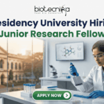 Presidency University Hiring Junior Research Fellow | Earn Upto ₹25,000 Salary! Presidency University Hiring Junior Research Fellow Earn Upto ₹25,000 Salary! A recruitment banner for Presidency University featuring the text 'Presidency University Hiring Junior Research Fellow' at the top, along with the 'biotecnika' logo. On the left side, the iconic colonial architecture of Presidency University's building is visible through a window, with a view of the sun setting. The right side features a female Indian scientist of a professional and focused appearance, wearing a lab coat, safety glasses, and blue gloves, meticulously using a pipette to fill a well-plate. She is surrounded by modern laboratory equipment, including a microscope and a monitor displaying molecular structures. Floating icons of DNA double helices are subtly placed around the text area. An 'APPLY NOW' button is prominent at the bottom center.