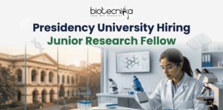 Presidency University Hiring Junior Research Fellow Earn Upto ₹25,000 Salary! A recruitment banner for Presidency University featuring the text 'Presidency University Hiring Junior Research Fellow' at the top, along with the 'biotecnika' logo. On the left side, the iconic colonial architecture of Presidency University's building is visible through a window, with a view of the sun setting. The right side features a female Indian scientist of a professional and focused appearance, wearing a lab coat, safety glasses, and blue gloves, meticulously using a pipette to fill a well-plate. She is surrounded by modern laboratory equipment, including a microscope and a monitor displaying molecular structures. Floating icons of DNA double helices are subtly placed around the text area. An 'APPLY NOW' button is prominent at the bottom center.