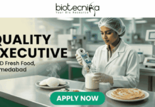 Quality Executive at iD Fresh Food Ahmedabad – Food Tech Graduates Apply Now! Quality Executive at iD Fresh Food Ahmedabad. A recruitment banner for the role of "QUALITY EXECUTIVE at iD Fresh Food, Ahmedabad". At the top center is the "biotecnika Your Bio Resource" logo. The main image features a focused South Asian woman in a professional lab coat and hairnet, working in a modern, well-lit food laboratory. She is using a hand-held digital analytical testing device on a sample in a glass beaker. In the foreground on a stainless steel counter are steaming plates of fresh Idlis with chutney and two Parottas. The text "QUALITY EXECUTIVE at iD Fresh Food, Ahmedabad" is on the left. A prominent green "APPLY NOW" button is at the bottom. The setting is clean and industrial, with green accents.