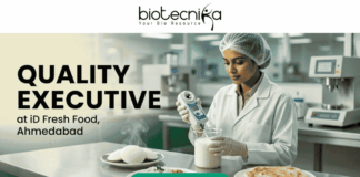 Quality Executive at iD Fresh Food Ahmedabad – Food Tech Graduates Apply Now! Quality Executive at iD Fresh Food Ahmedabad. A recruitment banner for the role of "QUALITY EXECUTIVE at iD Fresh Food, Ahmedabad". At the top center is the "biotecnika Your Bio Resource" logo. The main image features a focused South Asian woman in a professional lab coat and hairnet, working in a modern, well-lit food laboratory. She is using a hand-held digital analytical testing device on a sample in a glass beaker. In the foreground on a stainless steel counter are steaming plates of fresh Idlis with chutney and two Parottas. The text "QUALITY EXECUTIVE at iD Fresh Food, Ahmedabad" is on the left. A prominent green "APPLY NOW" button is at the bottom. The setting is clean and industrial, with green accents.