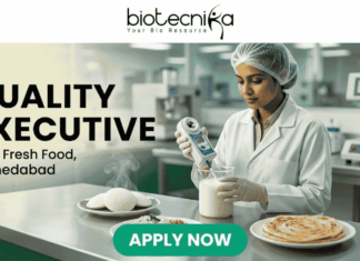Quality Executive at iD Fresh Food Ahmedabad – Food Tech Graduates Apply Now! Quality Executive at iD Fresh Food Ahmedabad. A recruitment banner for the role of "QUALITY EXECUTIVE at iD Fresh Food, Ahmedabad". At the top center is the "biotecnika Your Bio Resource" logo. The main image features a focused South Asian woman in a professional lab coat and hairnet, working in a modern, well-lit food laboratory. She is using a hand-held digital analytical testing device on a sample in a glass beaker. In the foreground on a stainless steel counter are steaming plates of fresh Idlis with chutney and two Parottas. The text "QUALITY EXECUTIVE at iD Fresh Food, Ahmedabad" is on the left. A prominent green "APPLY NOW" button is at the bottom. The setting is clean and industrial, with green accents.