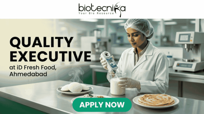 Quality Executive at iD Fresh Food Ahmedabad Quality Executive at iD Fresh Food Ahmedabad. A recruitment banner for the role of "QUALITY EXECUTIVE at iD Fresh Food, Ahmedabad". At the top center is the "biotecnika Your Bio Resource" logo. The main image features a focused South Asian woman in a professional lab coat and hairnet, working in a modern, well-lit food laboratory. She is using a hand-held digital analytical testing device on a sample in a glass beaker. In the foreground on a stainless steel counter are steaming plates of fresh Idlis with chutney and two Parottas. The text "QUALITY EXECUTIVE at iD Fresh Food, Ahmedabad" is on the left. A prominent green "APPLY NOW" button is at the bottom. The setting is clean and industrial, with green accents.