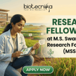 Research Fellow Job MSSRF | Botany Jobs India 2026. A job advertisement for 'RESEARCH FELLOW JOBS' at M.S. Swaminathan Research Foundation (MSSRF), in collaboration with biotecnika. The biotecnika logo is at the top. The image features a young woman scientist in a lab coat (with an MSSRF patch) kneeling in an experimental agricultural field (paddy field) and using a tablet that displays data graphs. The modern MSSRF facility, with its sign clearly visible, is in the background. The text 'RESEARCH FELLOW JOBS' is large and prominent. Small leaf and DNA icons are included. At the bottom, a green button reads 'APPLY NOW'.