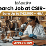 Research Job at CSIR-CLRI | Earn Up to ₹42,000 + HRA | Apply Before Deadline Research Job at CSIR-CLRI Earn Up to ₹42,000 + HRA. A promotional banner advertisement from Biotecnika, titled "Research Job at CSIR-CLRI", featuring a prominent gold banner with text that reads: "Earn Up to ₹42,000 + HRA". The main image shows a female scientist in a lab coat, safety glasses, and blue gloves, using a stereo microscope to analyze a textured material sample (likely a leather sample, given CLRI's focus). She is at a laboratory bench equipped with other microscopes, glass apparatus with samples, chemical bottles, and a computer monitor showing data. In the background are larger industrial lab machines. The Biotecnika "Your Bio Resource" logo is at the top center. At the bottom, a red button with white text reads: "APPLY NOW".