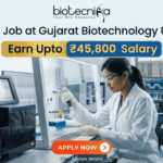 Research Job at Gujarat Biotechnology University | Earn Upto ₹45,800 Salary Research Job at Gujarat Biotechnology University ₹45,800 Salary. A promotional graphic for a Research Job at Gujarat Biotechnology University, featuring the 'biotecnika - Your Bio Resource' logo at the top. The main headline reads 'Research Job at Gujarat Biotechnology University' with prominent boxes below highlighting 'Earn Upto ₹45,800 Salary'. The background is a photograph of a female researcher in a white lab coat, gloves, and safety glasses, actively pipetting a multi-well plate within a modern, well-equipped genetics laboratory. Sophisticated sequencing equipment and monitors displaying DNA chromatograms and data are visible, along with a view of a university building through a window. A large, red 'APPLY NOW' button is at the bottom center with the text 'Click to view details'.