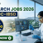 Research Jobs at BRIC-IBSD Walk-In Interview for Life Science Candidates. A professional recruitment banner for biotecnika advertising "Research Jobs 2026 at BRIC-IBSD." The top left features the biotecnika logo, and the headline text is prominent in blue and green. A circular blue callout on the left highlights the compensation: "Earn Upto ₹58,000 + HRA/ Month." On the right, a focused female scientist of professional and realistic appearance, wearing a white lab coat and safety glasses, is seated at a modern lab desk, using a pipette to fill a microplate. In the background, computer monitors display complex data including molecular structures and "Bioresource mapping" on a world map. Through a large window, a scenic modern building, perhaps the BRIC-IBSD institute, is visible in a lush environment. Two hexagonal icons on the far right show a network diagram over fresh produce and a sustainability tree icon. A prominent green "APPLY NOW" button with an arrow is at the bottom left.
