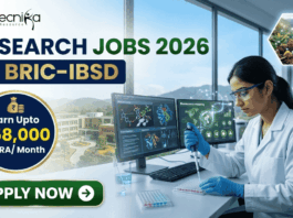 Research Jobs at BRIC-IBSD Walk-In Interview for Life Science Candidates. A professional recruitment banner for biotecnika advertising "Research Jobs 2026 at BRIC-IBSD." The top left features the biotecnika logo, and the headline text is prominent in blue and green. A circular blue callout on the left highlights the compensation: "Earn Upto ₹58,000 + HRA/ Month." On the right, a focused female scientist of professional and realistic appearance, wearing a white lab coat and safety glasses, is seated at a modern lab desk, using a pipette to fill a microplate. In the background, computer monitors display complex data including molecular structures and "Bioresource mapping" on a world map. Through a large window, a scenic modern building, perhaps the BRIC-IBSD institute, is visible in a lush environment. Two hexagonal icons on the far right show a network diagram over fresh produce and a sustainability tree icon. A prominent green "APPLY NOW" button with an arrow is at the bottom left.
