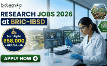 Research Jobs at BRIC-IBSD | Walk-In Interview for Life Science Candidates Research Jobs at BRIC-IBSD Walk-In Interview for Life Science Candidates. A professional recruitment banner for biotecnika advertising "Research Jobs 2026 at BRIC-IBSD." The top left features the biotecnika logo, and the headline text is prominent in blue and green. A circular blue callout on the left highlights the compensation: "Earn Upto ₹58,000 + HRA/ Month." On the right, a focused female scientist of professional and realistic appearance, wearing a white lab coat and safety glasses, is seated at a modern lab desk, using a pipette to fill a microplate. In the background, computer monitors display complex data including molecular structures and "Bioresource mapping" on a world map. Through a large window, a scenic modern building, perhaps the BRIC-IBSD institute, is visible in a lush environment. Two hexagonal icons on the far right show a network diagram over fresh produce and a sustainability tree icon. A prominent green "APPLY NOW" button with an arrow is at the bottom left.