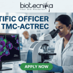 Scientific Officer Job at TMC-ACTREC. A job advertisement banner from biotecnika. At the top, the "biotecnika Your Bio Resource" logo with a stylized figure is displayed above the text 'SCIENTIFIC OFFICER JOB at TMC-ACTREC'. The main image shows a female scientist of South Asian appearance in a lab coat, looking through a complex multi-headed microscope. To her left, a computer monitor displays a vibrant, multi-colored confocal micrograph of stained cells with a scale bar, showcasing high-resolution cell imaging. Other researchers are working at distant lab benches in the background. Below the monitor, a text line reads, 'Explore groundbreaking oncology research in a world-class institute.' At the bottom center is a green 'APPLY NOW' button. The overall setting is a modern biological laboratory with various glass labware and equipment.