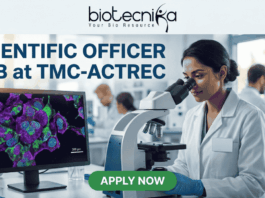 Scientific Officer Job at TMC-ACTREC. A job advertisement banner from biotecnika. At the top, the "biotecnika Your Bio Resource" logo with a stylized figure is displayed above the text 'SCIENTIFIC OFFICER JOB at TMC-ACTREC'. The main image shows a female scientist of South Asian appearance in a lab coat, looking through a complex multi-headed microscope. To her left, a computer monitor displays a vibrant, multi-colored confocal micrograph of stained cells with a scale bar, showcasing high-resolution cell imaging. Other researchers are working at distant lab benches in the background. Below the monitor, a text line reads, 'Explore groundbreaking oncology research in a world-class institute.' At the bottom center is a green 'APPLY NOW' button. The overall setting is a modern biological laboratory with various glass labware and equipment.