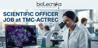 Scientific Officer Job at TMC-ACTREC. A job advertisement banner from biotecnika. At the top, the "biotecnika Your Bio Resource" logo with a stylized figure is displayed above the text 'SCIENTIFIC OFFICER JOB at TMC-ACTREC'. The main image shows a female scientist of South Asian appearance in a lab coat, looking through a complex multi-headed microscope. To her left, a computer monitor displays a vibrant, multi-colored confocal micrograph of stained cells with a scale bar, showcasing high-resolution cell imaging. Other researchers are working at distant lab benches in the background. Below the monitor, a text line reads, 'Explore groundbreaking oncology research in a world-class institute.' At the bottom center is a green 'APPLY NOW' button. The overall setting is a modern biological laboratory with various glass labware and equipment.