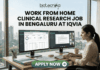 Work From Home Clinical Research Job in Bengaluru at IQVIA | Apply Now Work From Home Clinical Research Job in Bengaluru at IQVIA. A professional job advertisement banner for a "Work From Home Clinical Research Job in Bengaluru at IQVIA." The image features the Biotecnika logo at the top and a central headline in bold text. Below the text, a woman is shown working at a modern desk with a scenic city skyline view in the background. She is using a dual-monitor setup displaying clinical trial data management software and a video conference with a research team. A prominent green "Apply Now" button with a white arrow is located at the bottom center.