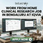 Work From Home Clinical Research Job in Bengaluru at IQVIA. A professional job advertisement banner for a "Work From Home Clinical Research Job in Bengaluru at IQVIA." The image features the Biotecnika logo at the top and a central headline in bold text. Below the text, a woman is shown working at a modern desk with a scenic city skyline view in the background. She is using a dual-monitor setup displaying clinical trial data management software and a video conference with a research team. A prominent green "Apply Now" button with a white arrow is located at the bottom center.