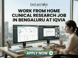 Work From Home Clinical Research Job in Bengaluru at IQVIA | Apply Now Work From Home Clinical Research Job in Bengaluru at IQVIA. A professional job advertisement banner for a "Work From Home Clinical Research Job in Bengaluru at IQVIA." The image features the Biotecnika logo at the top and a central headline in bold text. Below the text, a woman is shown working at a modern desk with a scenic city skyline view in the background. She is using a dual-monitor setup displaying clinical trial data management software and a video conference with a research team. A prominent green "Apply Now" button with a white arrow is located at the bottom center.