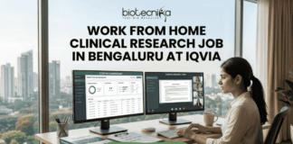 Work From Home Clinical Research Job in Bengaluru at IQVIA | Apply Now Work From Home Clinical Research Job in Bengaluru at IQVIA. A professional job advertisement banner for a "Work From Home Clinical Research Job in Bengaluru at IQVIA." The image features the Biotecnika logo at the top and a central headline in bold text. Below the text, a woman is shown working at a modern desk with a scenic city skyline view in the background. She is using a dual-monitor setup displaying clinical trial data management software and a video conference with a research team. A prominent green "Apply Now" button with a white arrow is located at the bottom center.