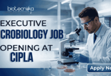 Executive Microbiology Job Opening at Cipla – MSc Candidates Apply Now Executive Microbiology Job Opening at Cipla – MSc Candidates Apply Now. A professional corporate recruitment banner for Cipla, sized 1920x1080 pixels with a blue gradient color scheme. On the left, bold text reads "EXECUTIVE MICROBIOLOGY JOB OPENING at Cipla," with "MICROBIOLOGY JOB" and "Cipla" prominently highlighted. On the right, a male microbiologist wearing a white lab coat, safety glasses, and blue gloves is focused on using a microscope in a clean, modern laboratory setting. The Cipla logo is positioned in the top right corner, and a distinct blue "Apply Now" button sits in the bottom right corner.