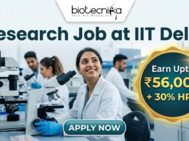 Research Job in Biomedical Engineering at IIT Delhi | Earn ₹56,000 per month. Corporate-style job featured image banner (1280x720) for "Research Job at IIT Delhi" featuring the Biotecnika logo at the top center. The background shows a realistic, modern laboratory where a smiling young Indian female researcher in a white lab coat sits with a microscope, while two colleagues work behind her. On the right, a prominent gold and teal circular badge highlights text reading "Earn Upto ₹56,000 + 30% HRA". A sleek teal "APPLY NOW" button is positioned at the bottom center.