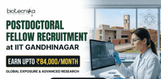 IIT Gandhinagar Postdoctoral Fellowship Earn Up to ₹84,000Month in Biomedical Research A corporate-style promotional banner for "Postdoctoral Fellow Recruitment at IIT Gandhinagar". The top left displays the Biotecnika logo, followed by bold text that reads "POSTDOCTORAL FELLOW RECRUITMENT at IIT GANDHINAGAR". A highlighted black and gold badge reads "EARN UPTO ₹84,000/MONTH", accompanied by the subtitle "GLOBAL EXPOSURE & ADVANCED RESEARCH". On the right side, a female researcher in a white lab coat, safety glasses, and blue gloves works at a computer monitor in a modern laboratory. Through the large window behind her, the iconic brick architecture of the IIT Gandhinagar campus is visible. A gold "APPLY NOW" button is positioned at the bottom center.