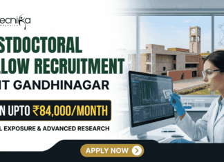 IIT Gandhinagar Postdoctoral Fellowship | Earn Up to ₹84,000/Month in Biomedical Research IIT Gandhinagar Postdoctoral Fellowship Earn Up to ₹84,000Month in Biomedical Research A corporate-style promotional banner for "Postdoctoral Fellow Recruitment at IIT Gandhinagar". The top left displays the Biotecnika logo, followed by bold text that reads "POSTDOCTORAL FELLOW RECRUITMENT at IIT GANDHINAGAR". A highlighted black and gold badge reads "EARN UPTO ₹84,000/MONTH", accompanied by the subtitle "GLOBAL EXPOSURE & ADVANCED RESEARCH". On the right side, a female researcher in a white lab coat, safety glasses, and blue gloves works at a computer monitor in a modern laboratory. Through the large window behind her, the iconic brick architecture of the IIT Gandhinagar campus is visible. A gold "APPLY NOW" button is positioned at the bottom center.