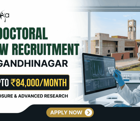 IIT Gandhinagar Postdoctoral Fellowship Earn Up to ₹84,000Month in Biomedical Research A corporate-style promotional banner for "Postdoctoral Fellow Recruitment at IIT Gandhinagar". The top left displays the Biotecnika logo, followed by bold text that reads "POSTDOCTORAL FELLOW RECRUITMENT at IIT GANDHINAGAR". A highlighted black and gold badge reads "EARN UPTO ₹84,000/MONTH", accompanied by the subtitle "GLOBAL EXPOSURE & ADVANCED RESEARCH". On the right side, a female researcher in a white lab coat, safety glasses, and blue gloves works at a computer monitor in a modern laboratory. Through the large window behind her, the iconic brick architecture of the IIT Gandhinagar campus is visible. A gold "APPLY NOW" button is positioned at the bottom center.