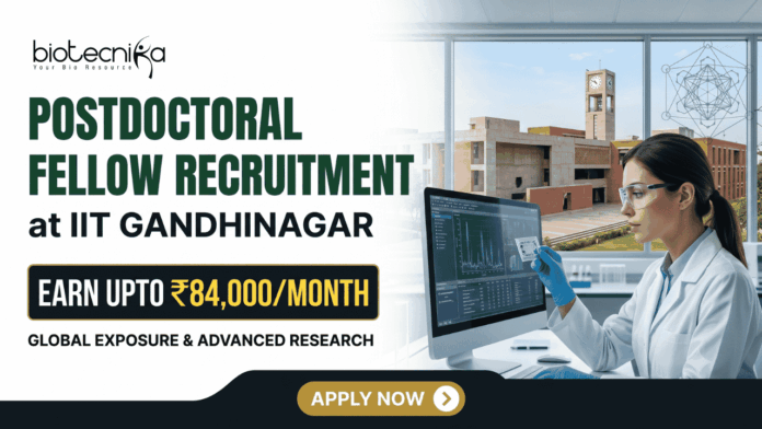 IIT Gandhinagar Postdoctoral Fellowship Earn Up to ₹84,000Month in Biomedical Research IIT Gandhinagar Postdoctoral Fellowship Earn Up to ₹84,000Month in Biomedical Research A corporate-style promotional banner for "Postdoctoral Fellow Recruitment at IIT Gandhinagar". The top left displays the Biotecnika logo, followed by bold text that reads "POSTDOCTORAL FELLOW RECRUITMENT at IIT GANDHINAGAR". A highlighted black and gold badge reads "EARN UPTO ₹84,000/MONTH", accompanied by the subtitle "GLOBAL EXPOSURE & ADVANCED RESEARCH". On the right side, a female researcher in a white lab coat, safety glasses, and blue gloves works at a computer monitor in a modern laboratory. Through the large window behind her, the iconic brick architecture of the IIT Gandhinagar campus is visible. A gold "APPLY NOW" button is positioned at the bottom center.