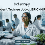 Student Trainee Job for Biotechnology & Life Science Students at BRIC-NIPGR Student Trainee Job for Biotechnology & Life Science Students at BRIC-NIPGR. A corporate-style job featured image banner (1280x720) for "Student Trainee Job at BRIC-NIPGR" featuring the Biotecnika CRO logo at the top center. The background showcases a realistic, high-tech plant biology research laboratory. A smiling young Indian female student trainee wearing a white lab coat, safety goggles, and blue gloves is using a pipette over a well plate. Trays of green plant seedlings are arranged on the lab counter alongside modern equipment, with other researchers working in the background. A clean, dark teal "APPLY NOW" button is centered at the bottom.