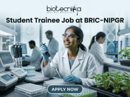 Student Trainee Job for Biotechnology & Life Science Students at BRIC-NIPGR. A corporate-style job featured image banner (1280x720) for "Student Trainee Job at BRIC-NIPGR" featuring the Biotecnika CRO logo at the top center. The background showcases a realistic, high-tech plant biology research laboratory. A smiling young Indian female student trainee wearing a white lab coat, safety goggles, and blue gloves is using a pipette over a well plate. Trays of green plant seedlings are arranged on the lab counter alongside modern equipment, with other researchers working in the background. A clean, dark teal "APPLY NOW" button is centered at the bottom.